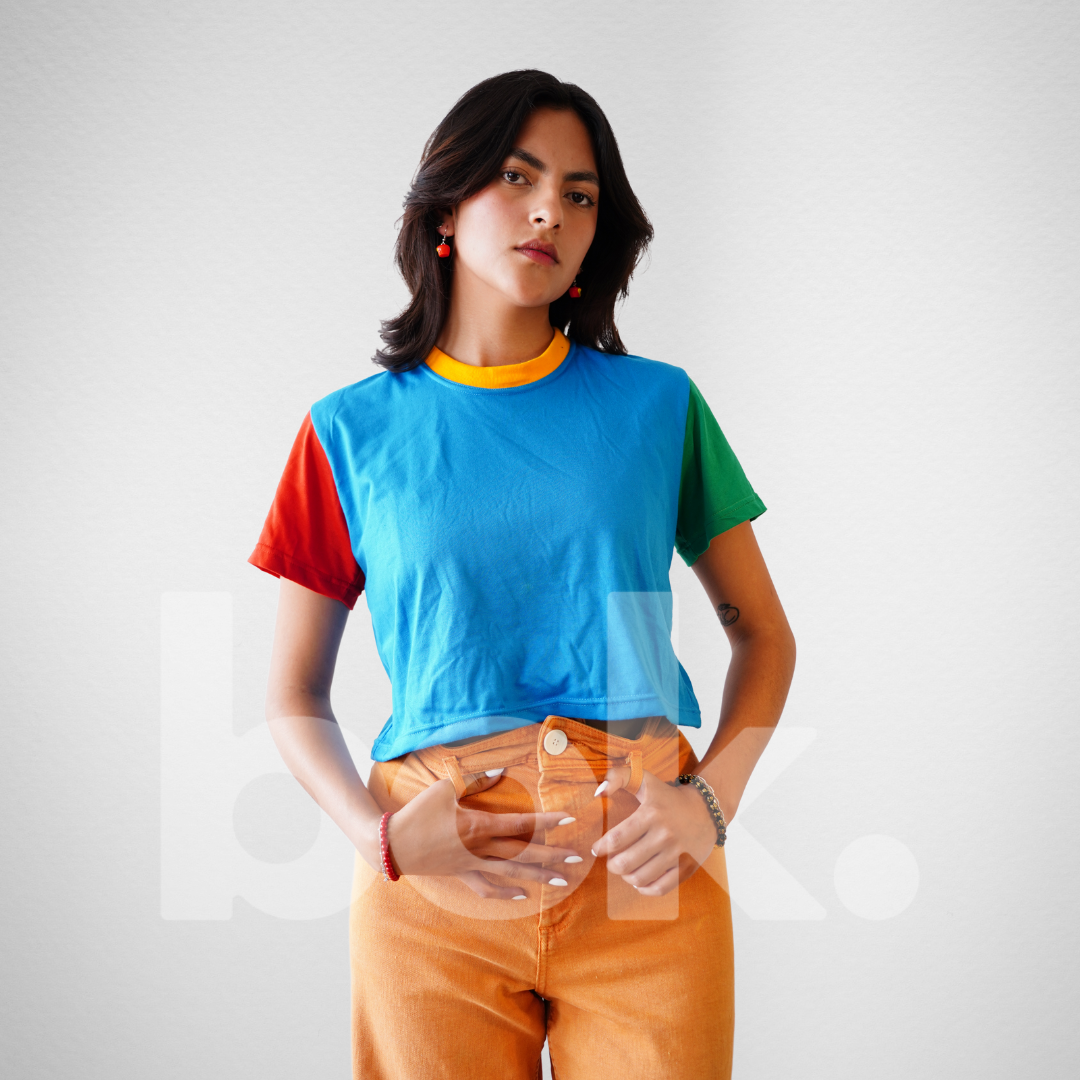 Modelo luciendo un crop top retro celeste con detalles en rojo, verde y amarillo, combinado con pantalones naranja para un look casual, fresco y vibrante.