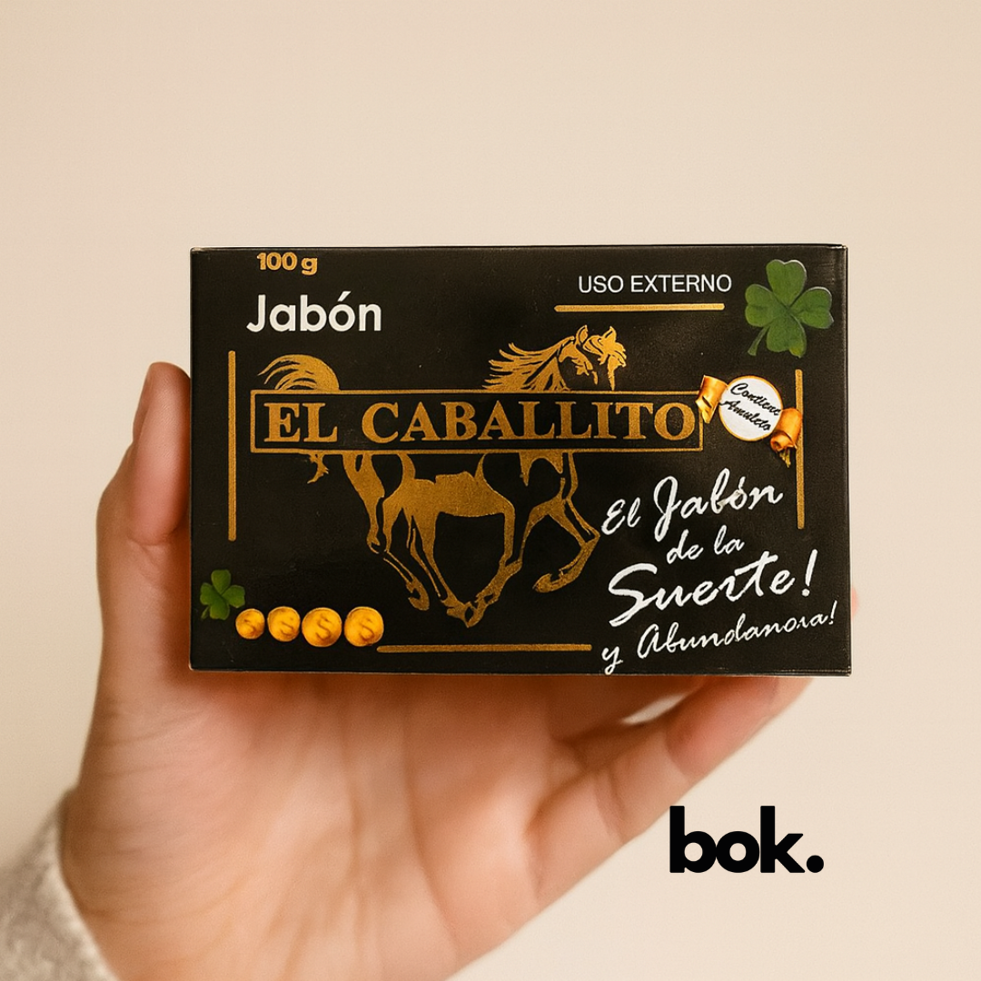 Jabón El Caballito artesanal BOK con amuleto, caja negra con diseño dorado de caballo y trébol de cuatro hojas, sostenido por una mano sobre fondo neutro, producto natural.