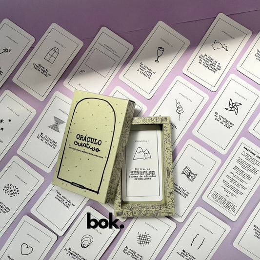 Caja morada del Oráculo Creativo by Vayeca abierta sobre una superficie lila. A su alrededor, varias cartas con frases e ilustraciones minimalistas. El mazo incluye 100 afirmaciones diseñadas para activar y fortalecer la creatividad.