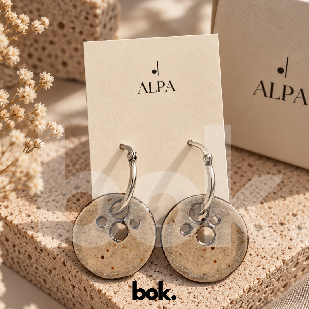 Aretes Luna de cerámica artesanal ALPA hechos a mano en Ecuador, en tono neutro con perforaciones inspiradas en cráteres y aro plateado, disponibles en BOK Concept Store Quito.