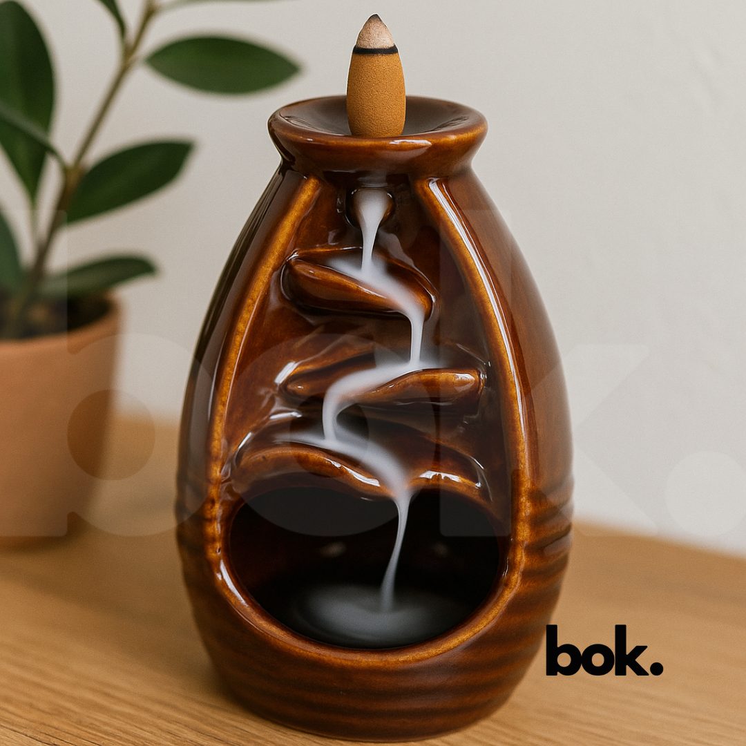 Porta conos de cerámica color café con cono de incienso encendido y humo cayendo en forma de cascada sobre superficie de madera, decoración artesanal disponible en BOK Concept Store.