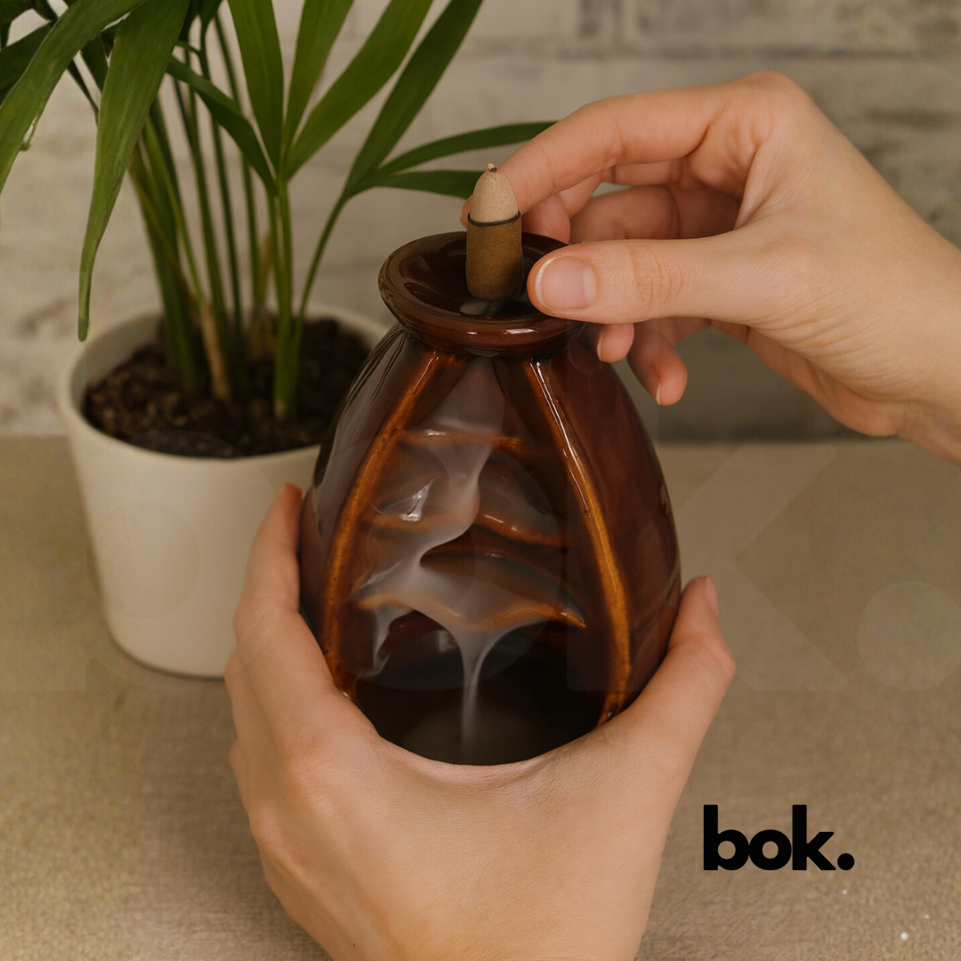 Porta conos de cerámica color café con cono de incienso encendido y humo cayendo en forma de cascada sobre superficie de madera, decoración artesanal disponible en BOK Concept Store.
