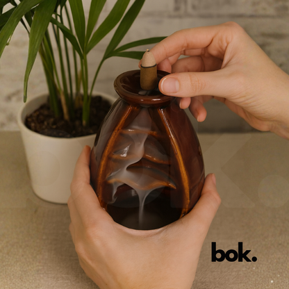 Porta conos de cerámica color café con cono de incienso encendido y humo cayendo en forma de cascada sobre superficie de madera, decoración artesanal disponible en BOK Concept Store.