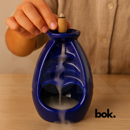 Porta conos de cerámica color azul con cono de incienso encendido, humo descendiendo en cascada y fondo cálido, producto artesanal decorativo de BOK Concept Store.