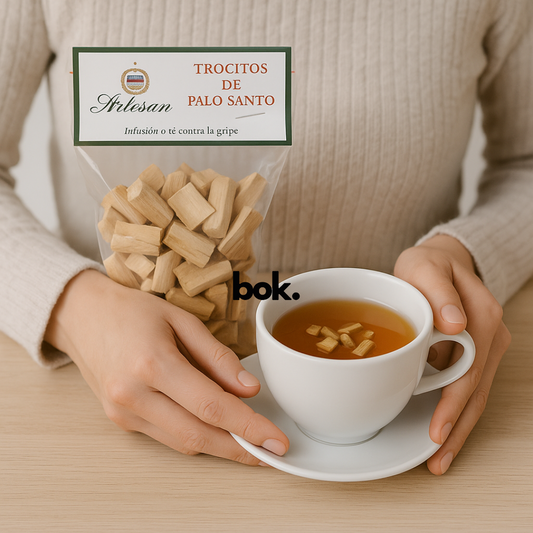 Persona disfrutando una taza de infusión de Palo Santo con trocitos pequeños, junto a bolsa artesanal de BOK Concept Store en mesa de madera clara.