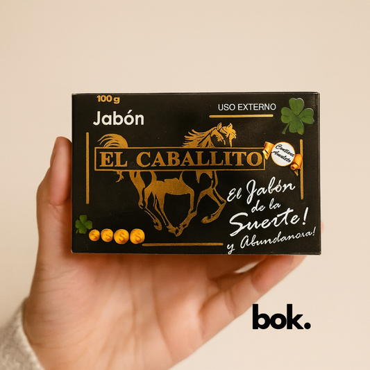 Jabón El Caballito artesanal BOK con amuleto, caja negra con diseño dorado de caballo y trébol de cuatro hojas, sostenido por una mano sobre fondo neutro, producto natural.