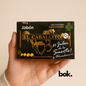 Jabón El Caballito artesanal BOK con amuleto, caja negra con diseño dorado de caballo y trébol de cuatro hojas, sostenido por una mano sobre fondo neutro, producto natural.