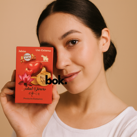 Jabón Salud Dinero Amor artesanal BOK con amuleto, en caja roja con diseño de corazones, monedas y flor de loto, sostenido por una mujer sobre fondo neutro, producto natural.