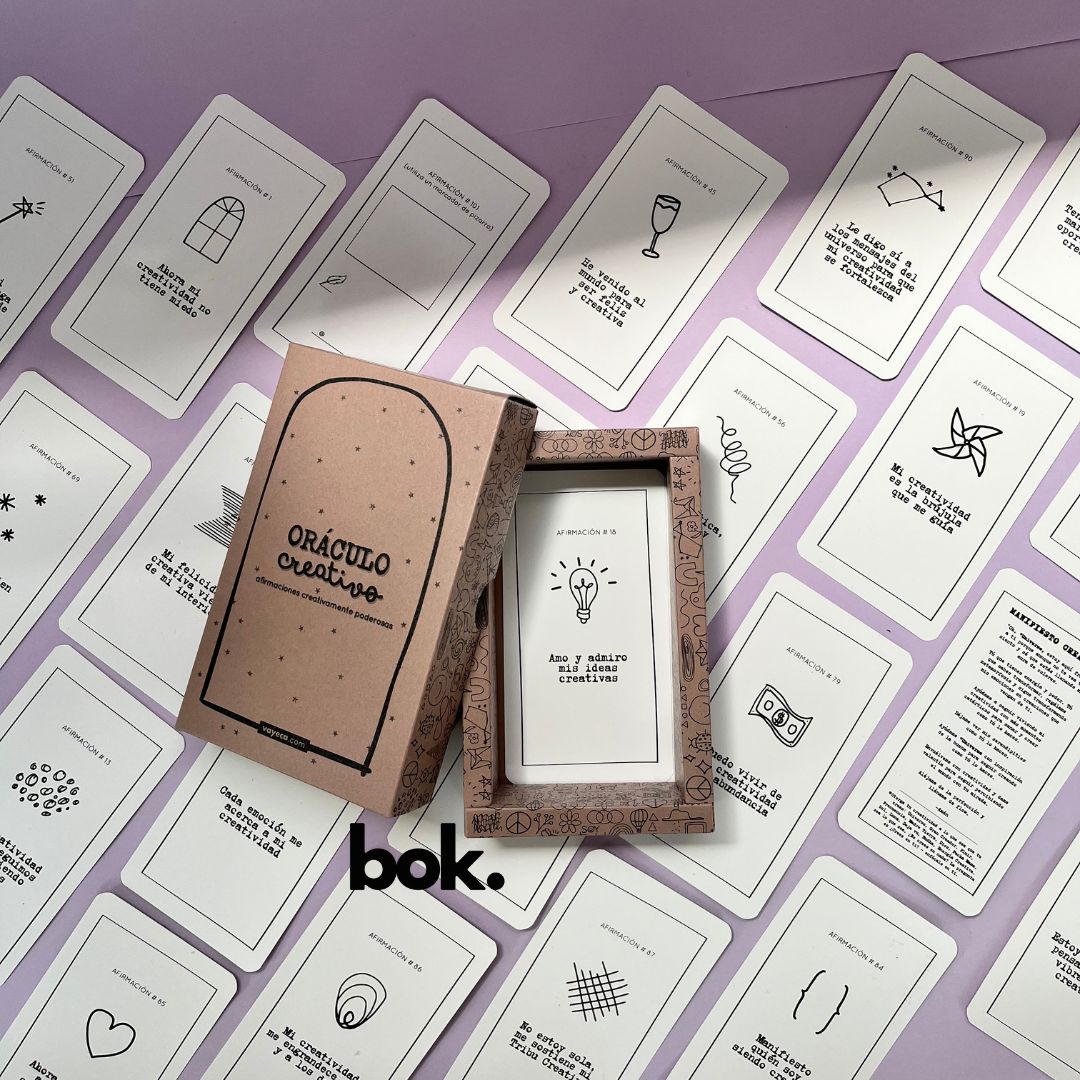 Caja morada del Oráculo Creativo by Vayeca abierta sobre una superficie lila. A su alrededor, varias cartas con frases e ilustraciones minimalistas. El mazo incluye 100 afirmaciones diseñadas para activar y fortalecer la creatividad.