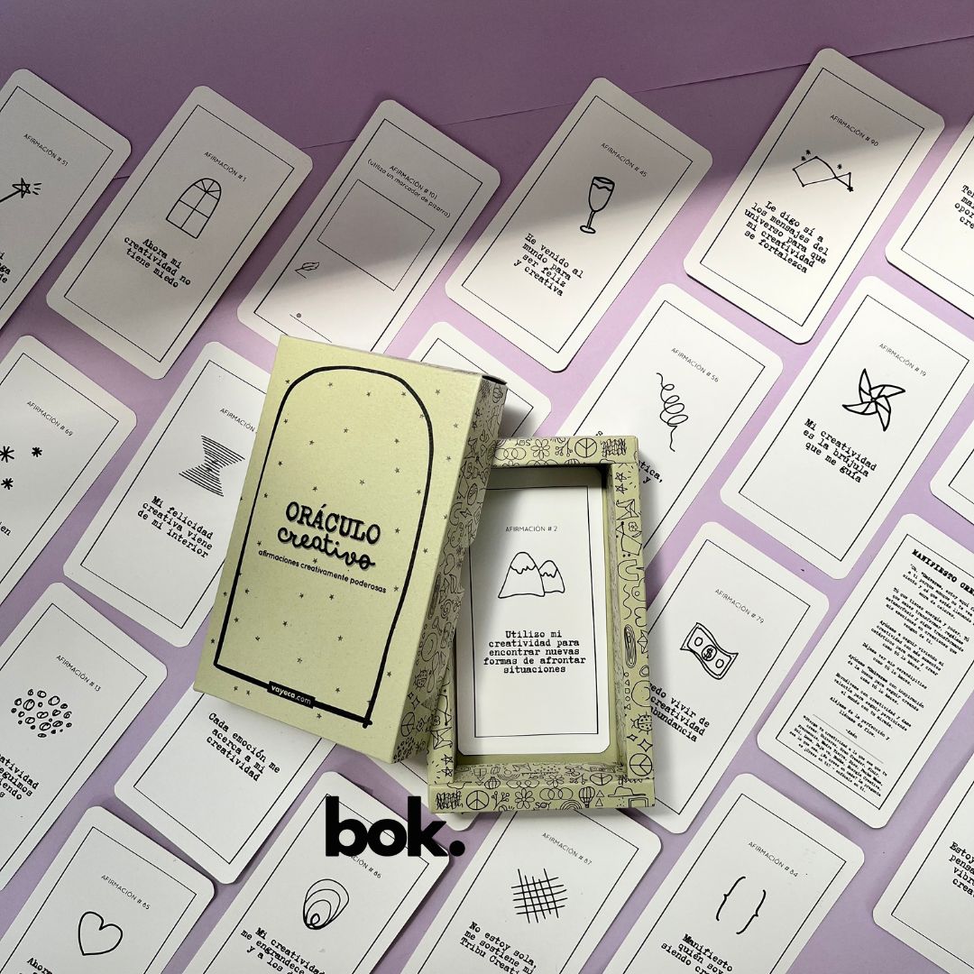 Caja morada del Oráculo Creativo by Vayeca abierta sobre una superficie lila. A su alrededor, varias cartas con frases e ilustraciones minimalistas. El mazo incluye 100 afirmaciones diseñadas para activar y fortalecer la creatividad.