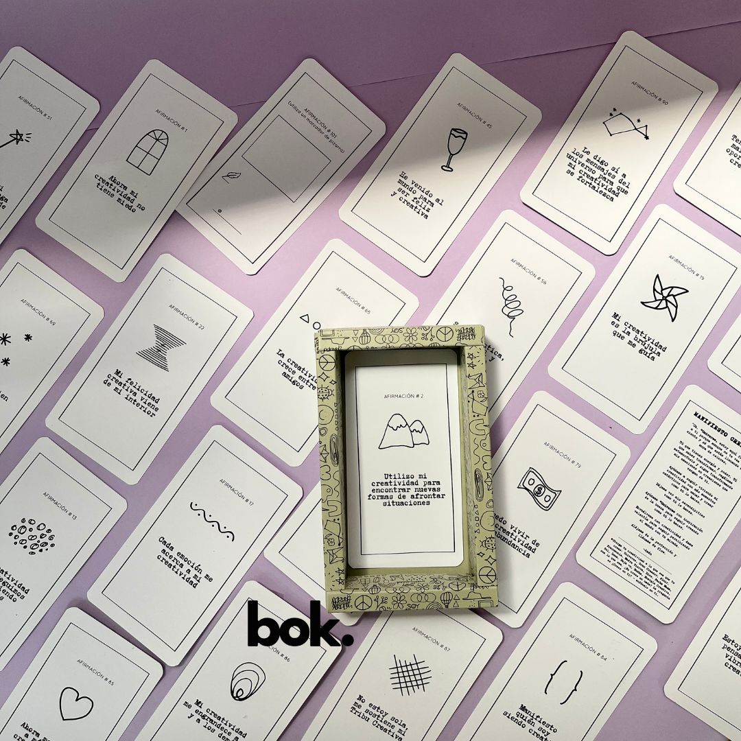 Caja morada del Oráculo Creativo by Vayeca abierta sobre una superficie lila. A su alrededor, varias cartas con frases e ilustraciones minimalistas. El mazo incluye 100 afirmaciones diseñadas para activar y fortalecer la creatividad.