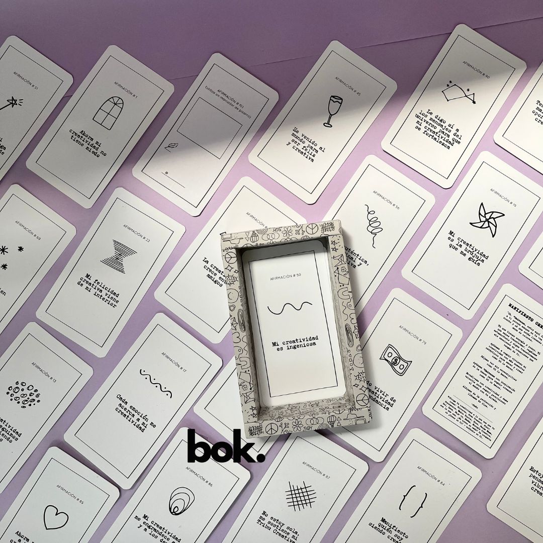 Caja morada del Oráculo Creativo by Vayeca abierta sobre una superficie lila. A su alrededor, varias cartas con frases e ilustraciones minimalistas. El mazo incluye 100 afirmaciones diseñadas para activar y fortalecer la creatividad.