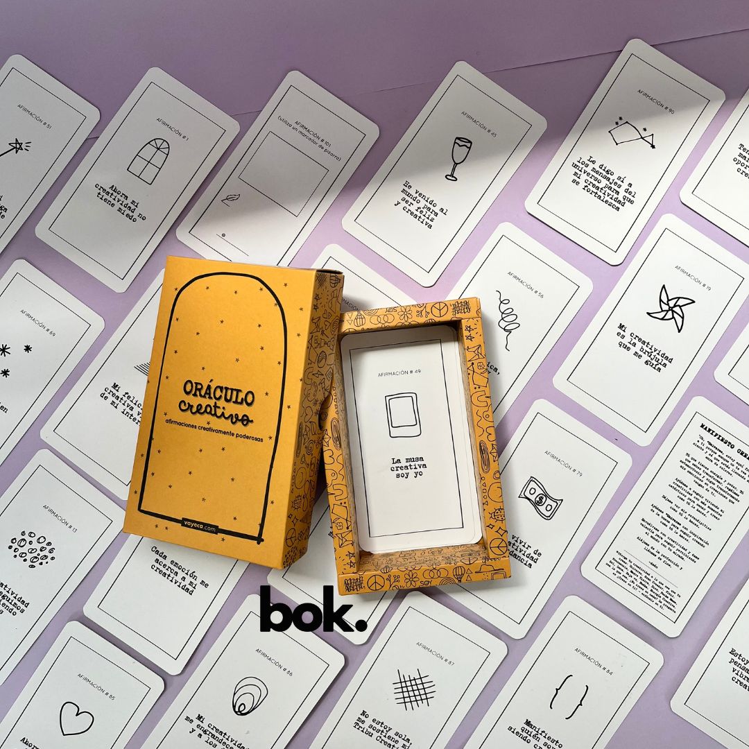 Caja morada del Oráculo Creativo by Vayeca abierta sobre una superficie lila. A su alrededor, varias cartas con frases e ilustraciones minimalistas. El mazo incluye 100 afirmaciones diseñadas para activar y fortalecer la creatividad.