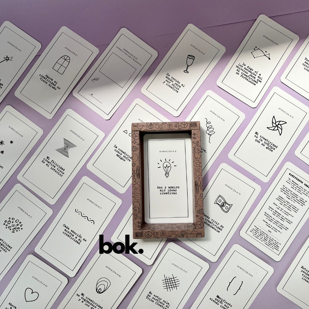 Caja morada del Oráculo Creativo by Vayeca abierta sobre una superficie lila. A su alrededor, varias cartas con frases e ilustraciones minimalistas. El mazo incluye 100 afirmaciones diseñadas para activar y fortalecer la creatividad.