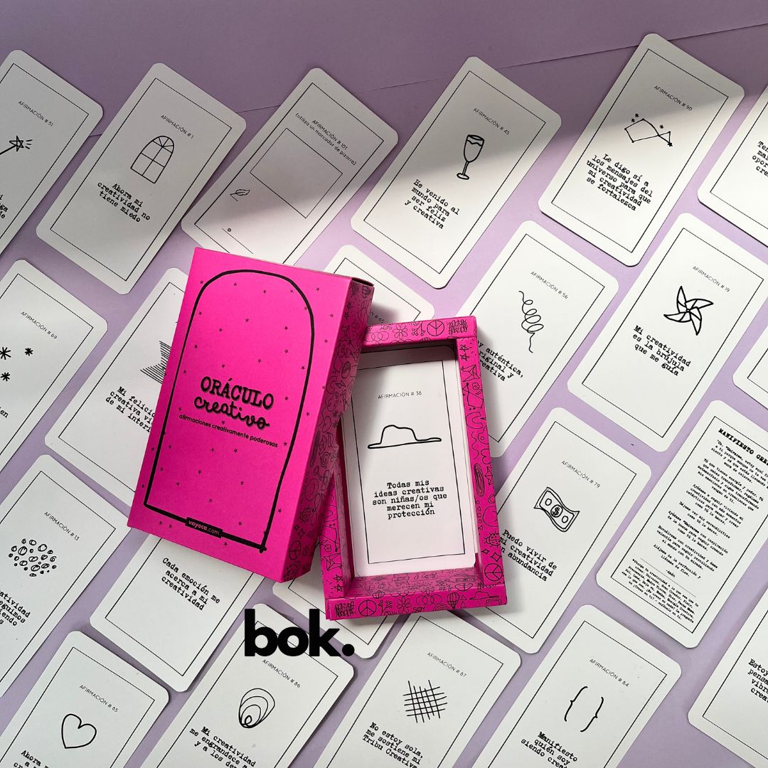 Caja morada del Oráculo Creativo by Vayeca abierta sobre una superficie lila. A su alrededor, varias cartas con frases e ilustraciones minimalistas. El mazo incluye 100 afirmaciones diseñadas para activar y fortalecer la creatividad.