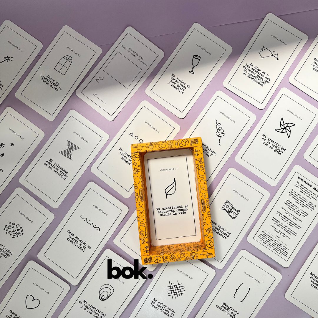 Caja morada del Oráculo Creativo by Vayeca abierta sobre una superficie lila. A su alrededor, varias cartas con frases e ilustraciones minimalistas. El mazo incluye 100 afirmaciones diseñadas para activar y fortalecer la creatividad.