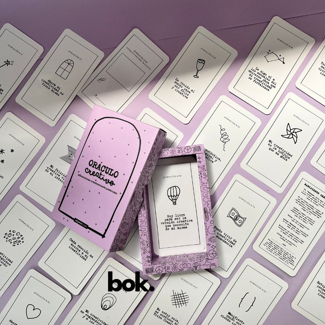 Caja morada del Oráculo Creativo by Vayeca abierta sobre una superficie lila. A su alrededor, varias cartas con frases e ilustraciones minimalistas. El mazo incluye 100 afirmaciones diseñadas para activar y fortalecer la creatividad.
