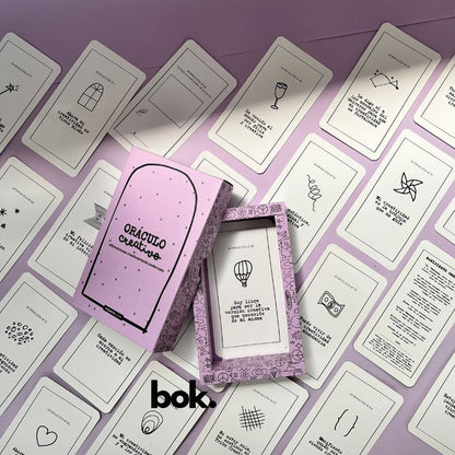 Caja morada del Oráculo Creativo by Vayeca abierta sobre una superficie lila. A su alrededor, varias cartas con frases e ilustraciones minimalistas. El mazo incluye 100 afirmaciones diseñadas para activar y fortalecer la creatividad.
