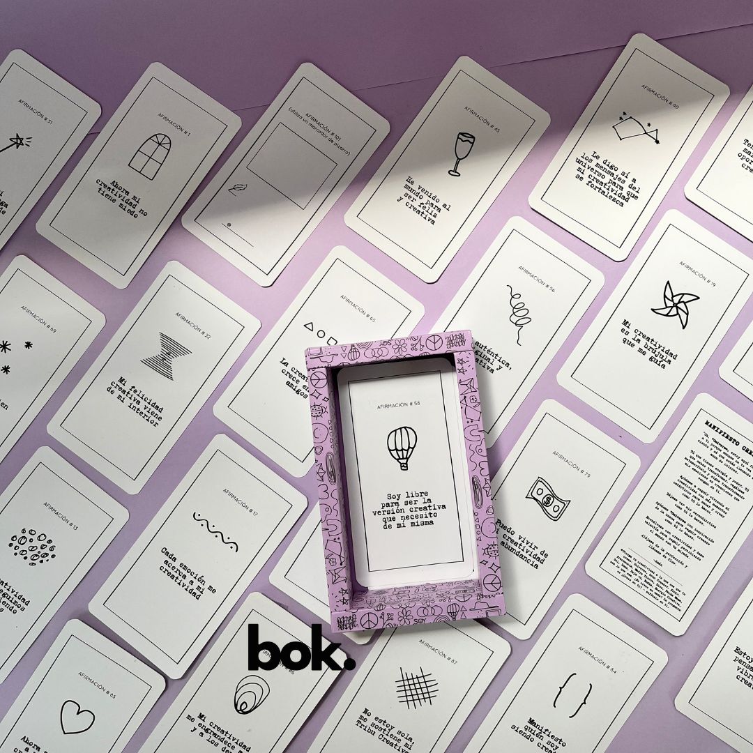 Caja morada del Oráculo Creativo by Vayeca abierta sobre una superficie lila. A su alrededor, varias cartas con frases e ilustraciones minimalistas. El mazo incluye 100 afirmaciones diseñadas para activar y fortalecer la creatividad.