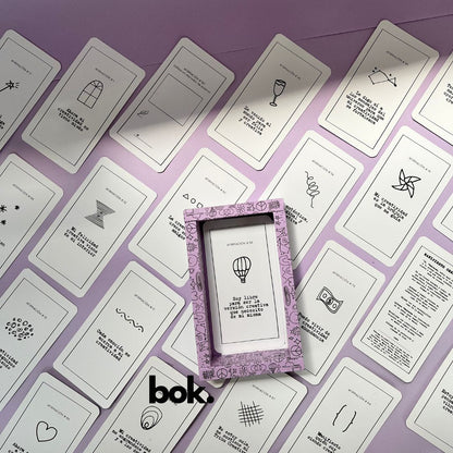 Caja morada del Oráculo Creativo by Vayeca abierta sobre una superficie lila. A su alrededor, varias cartas con frases e ilustraciones minimalistas. El mazo incluye 100 afirmaciones diseñadas para activar y fortalecer la creatividad.