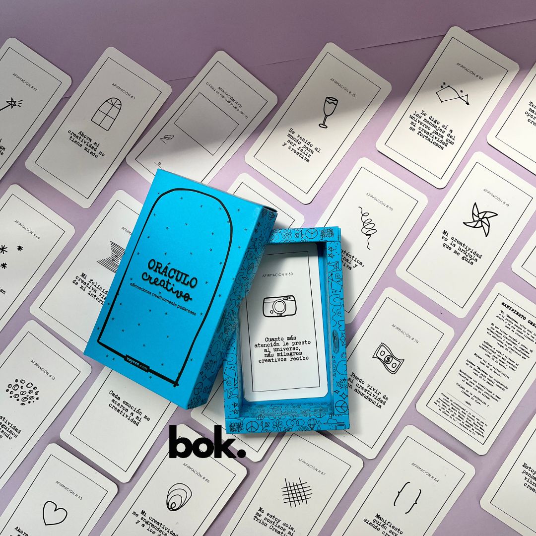 Caja morada del Oráculo Creativo by Vayeca abierta sobre una superficie lila. A su alrededor, varias cartas con frases e ilustraciones minimalistas. El mazo incluye 100 afirmaciones diseñadas para activar y fortalecer la creatividad.