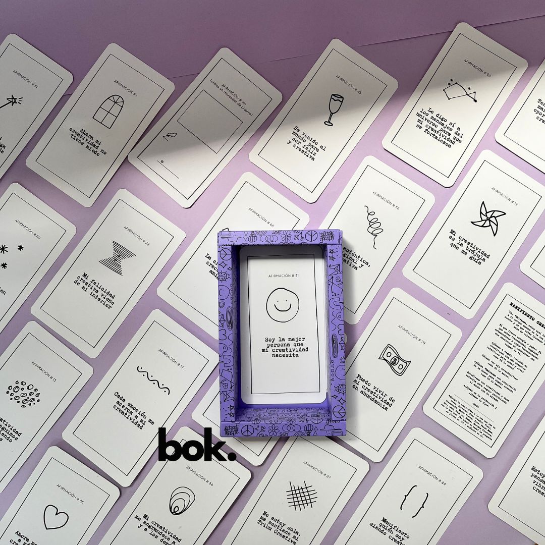 Caja morada del Oráculo Creativo by Vayeca abierta sobre una superficie lila. A su alrededor, varias cartas con frases e ilustraciones minimalistas. El mazo incluye 100 afirmaciones diseñadas para activar y fortalecer la creatividad.