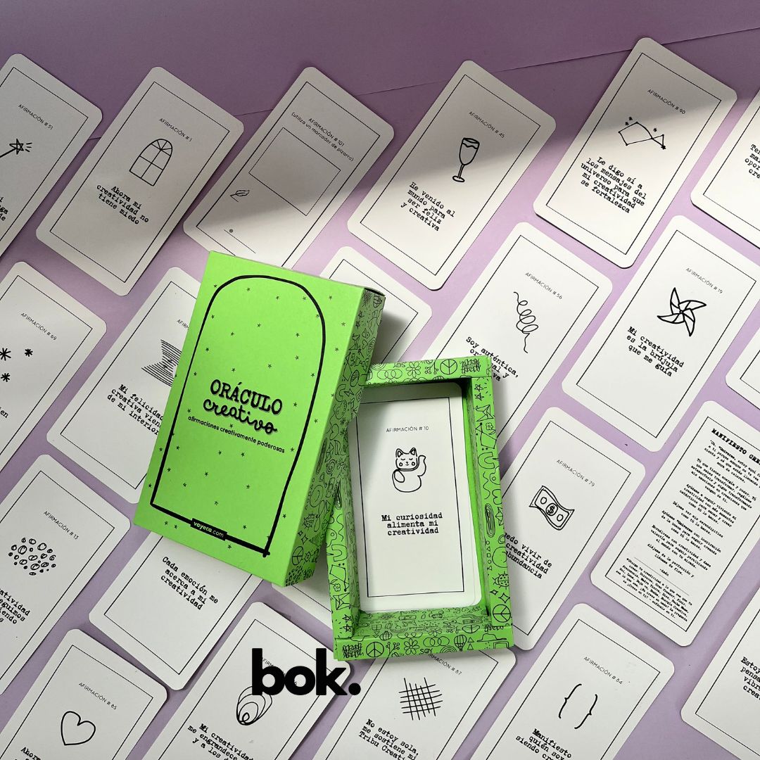 Caja morada del Oráculo Creativo by Vayeca abierta sobre una superficie lila. A su alrededor, varias cartas con frases e ilustraciones minimalistas. El mazo incluye 100 afirmaciones diseñadas para activar y fortalecer la creatividad.