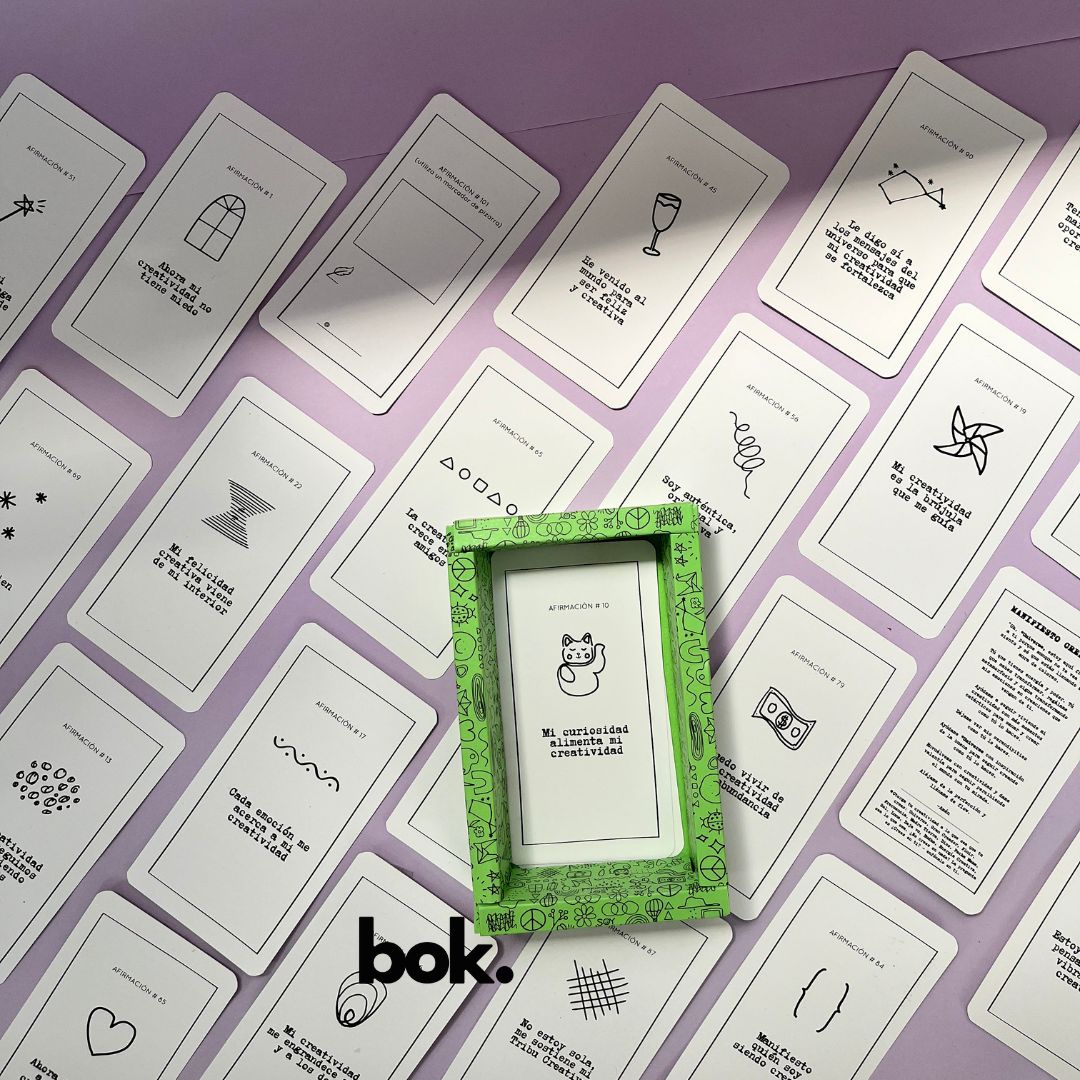 Caja morada del Oráculo Creativo by Vayeca abierta sobre una superficie lila. A su alrededor, varias cartas con frases e ilustraciones minimalistas. El mazo incluye 100 afirmaciones diseñadas para activar y fortalecer la creatividad.