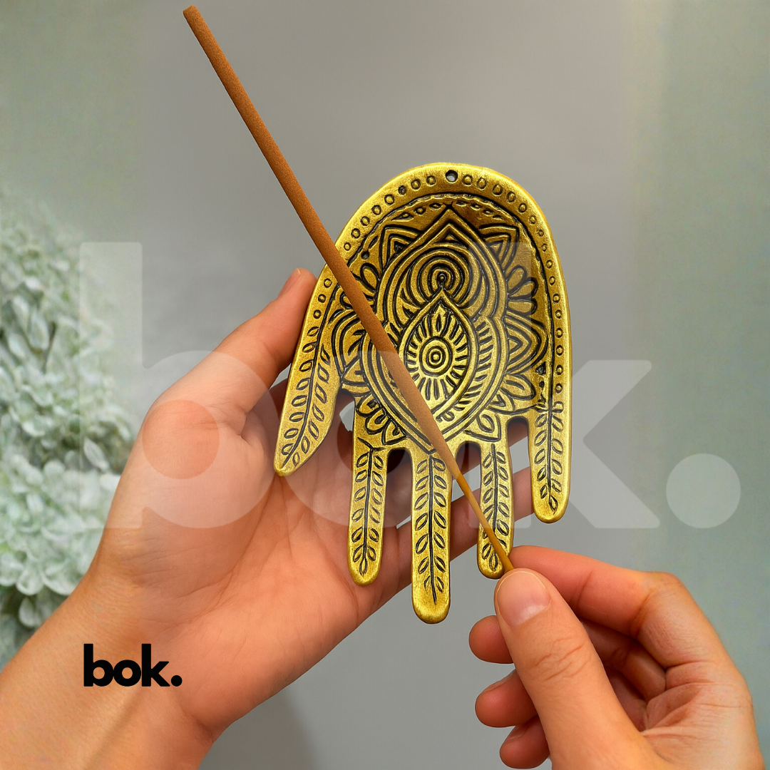 Persona sosteniendo un incienso para colocarlo en un porta inciensos dorado con forma de mano hindú, en un ambiente elegante y luminoso.