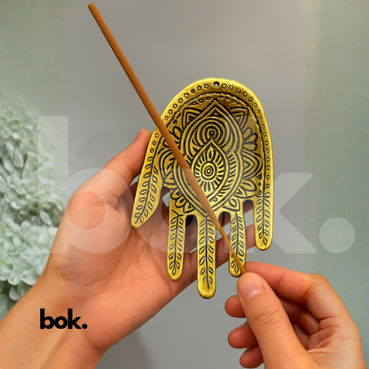 Persona sosteniendo un incienso para colocarlo en un porta inciensos dorado con forma de mano hindú, en un ambiente elegante y luminoso.