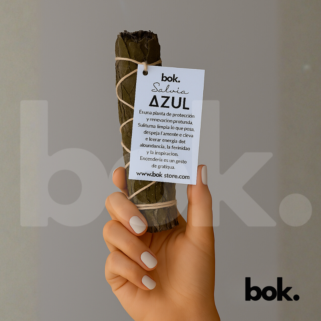 Sahumerio de Salvia Azul de BOK sostenido en una mano, con humo elevándose y etiqueta visible. Fondo gris minimalista y luz cálida que resaltan su textura natural y energía protectora.