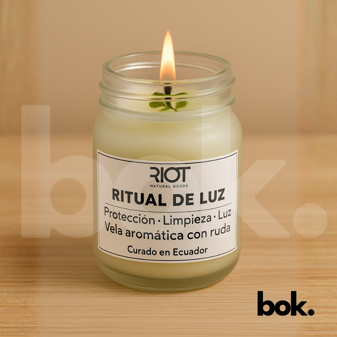 Vela Ritual de Luz encendida sobre una superficie de lino beige, con su etiqueta visible y una ramita de ruda dentro del frasco. Imagen minimalista y cálida que representa protección y serenidad.