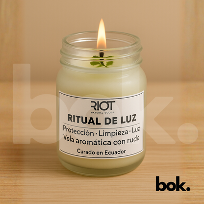 Vela Ritual de Luz encendida sobre una superficie de lino beige, con su etiqueta visible y una ramita de ruda dentro del frasco. Imagen minimalista y cálida que representa protección y serenidad.